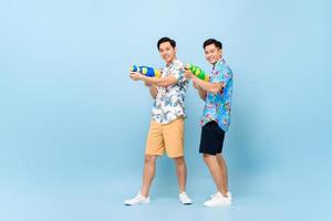 Smilng happy Asian male friends playing with water guns in blue isolated background for Songkran festival in Thailand and southeast Asia photo