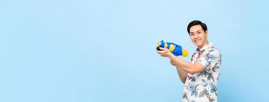 Smiling handsome Asian man playing with water gun isolated on banner blue background with copy space for Songkran festival in Thailand and southeast Asia photo