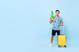 Smiling handsome Asian tourist man traveling with water gun and baggage during Songkran festival studio shot blue background with copy space photo