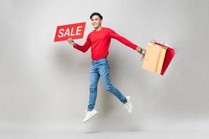 Surprised Asian man holding shopping bags and red sale sign jumping in isolated light gray studio background for Chinese new year sale concept