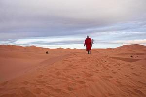 Man in traditional clothes with sandboard walking on sand dunes against sky photo