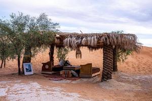 Sofa with cushions under hut on sand dunes in desert against cloudy sky photo