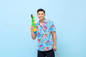 hombre asiático guapo sonriente jugando con pistola de agua aislado en el fondo azul del estudio para el festival songkran en tailandia y el sudeste asiático foto