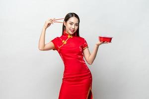 Pretty Asian woman in traditional red Chinese qipao dress holding plastic food box and chopsticks in studio isolated gray background photo