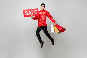 Surprised Asian man in traditional costume holding bags and red sale sign jumping in isolated light gray background for Chinese new year shopping concept