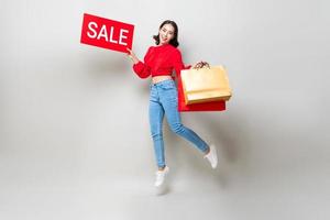 Happy Asian woman holding shopping bags and red sale sign jumping in isolated light gray studio background for Chinese new year sale concept