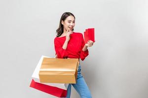 hermosa mujer asiática sonriente sosteniendo bolsas de compras y sobres rojos aislados en un estudio gris para el concepto de venta de año nuevo chino foto