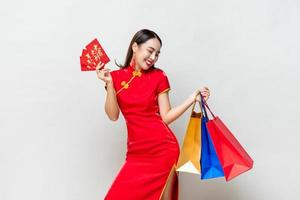 Happy Asian woman in traditional dress holding bags and red envelopes in isolated light gray studio background for Chinese new year shopping concept, text means great luck great profit