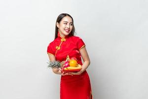 Young Asian woman in traditinal red cheongsam dress holding lucky fruits for Chinese new year in light gray isolated studio background