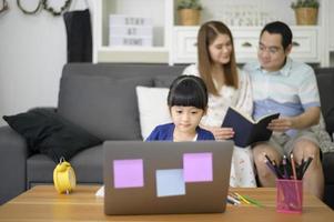 la feliz hija asiática está usando una computadora portátil para estudiar en línea a través de Internet mientras los padres se sientan en el sofá en casa. concepto de aprendizaje electrónico foto