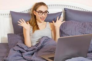 Emotional Happy Woman With Glasses Lies On The Bed With Laptop photo