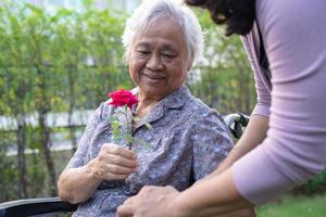 cuidadora hija abrazo y ayuda a anciana asiática mayor o anciana sosteniendo una rosa roja en silla de ruedas en el parque. foto