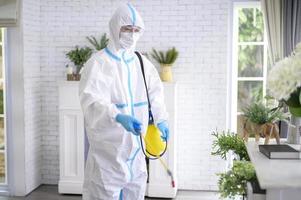 A medical staff in PPE suit is using disinfectant spray in living room, Covid-19 protection , disinfection concept . photo