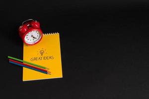 alarm clock with notepad and colored pencils on black background, isolated. back to school. great ideas photo