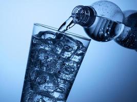 verter agua de una botella de plástico en un vaso con hielo sobre un fondo azul claro foto