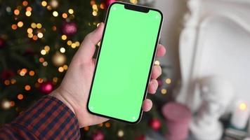 hombre que sostiene un teléfono inteligente moderno con cromakey de pantalla verde cerca de las luces del árbol de Navidad en el fondo video