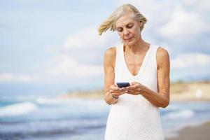 Senior woman walking on the beach using a smartphone.