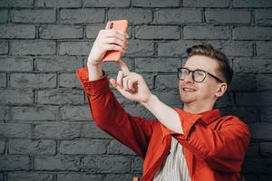 Young joyful man in red shirt and glasses taking selfie photo on cellphone on a background of black brick wall. Copy, empty space for text