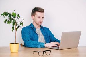 Young man in shirt and glasses working on laptop computer sitting at the table on a white background. Freelance work concept. Copy, empty space for text photo