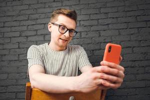joven con anteojos mirando el teléfono inteligente mientras se sienta en una silla sobre un fondo de pared de ladrillo negro. copiar, espacio vacío para texto