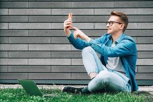 Man with glasses sitting on green grass and looking at smartphone on a gray wall background photo