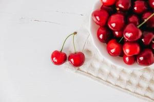 Fresh juicy red cherries in a white plate on the white wooden background. Top view. Copy, empty space for text