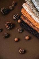 Stack of knitted material from threads of brown, orange, gray colors with pine cones a brown background. Top view. Copy, empty space for text photo