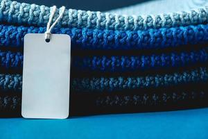 Stack of knitted material from threads of dark blue, light blue, gray colors with blank Price Tag on a blue background. Copy, empty space for text photo