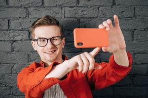 Young joyful man in red shirt and glasses taking selfie photo on cellphone on a background of black brick wall. Copy, empty space for text