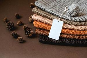 Stack of knitted material from threads of brown, orange, gray colors with an empty price tag on a brown background. Copy, empty space for text photo