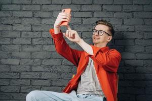 Young joyful man in red shirt and glasses taking selfie photo on cellphone on a background of black brick wall. Copy, empty space for text
