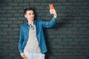 Young man in blue shirt and glasses taking selfie photo on cellphone on a background of black brick wall. Copy, empty space for text