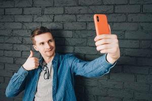 Young joyful man in blue shirt and glasses taking selfie photo on cellphone on a background of black brick wall. Copy, empty space for text