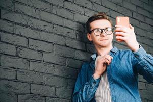 Young man in blue shirt and glasses taking selfie photo on cellphone on a background of black brick wall. Copy, empty space for text