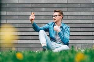 Man with glasses sitting on green grass and looking at smartphone on a gray wall background photo