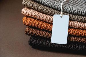 Stack of knitted material from threads of brown, orange, gray colors with an empty price tag on a brown background. Copy, empty space for text photo