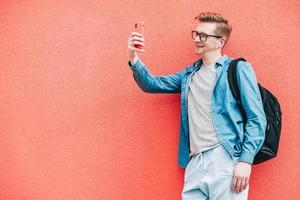 Man in shirt, jeans and glasses carrying a backpack and looking at smartphone while standing on a pink background photo