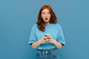 mujer pelirroja sorprendida en camiseta usando teléfono móvil