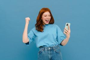 mujer pelirroja emocionada en camiseta gesticulando y usando teléfono celular foto