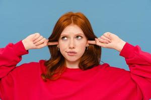 Perplexed ginger-haired woman wearing red sweater plugging her ears photo