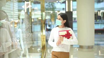 mujer con máscara protectora sosteniendo una caja de regalo en un centro comercial, comprando bajo la pandemia de covid-19, acción de gracias y concepto de navidad. video