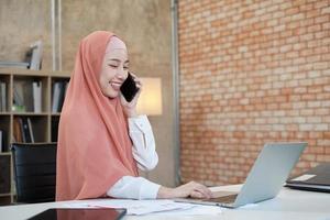 hermosa mujer de negocios que persona de sexo femenino islámico está hablando por la comunicación de su teléfono móvil y sonriendo feliz porque orden en línea de Internet en la computadora portátil, estilo de vida moderno, ocupación de comercio electrónico. foto