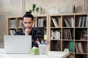 Young male worker of Asian ethnicity uses laptop to do creative work on white desk in front of bookshelf of a casual workplace, startup business person, and online e-commerce occupation. photo