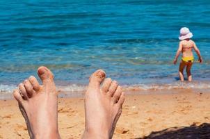 female feet in the sand by the sea and a child in a panama hat by the coast photo