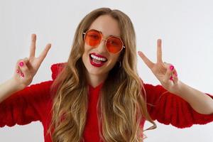 Young woman in red hoodie sweatshirt isolated on white background. Fashion and style concept. Peace sign gesture. Happy beautiful girl in red sunglasses. Make-up and beauty. Glamour model. Kiss face photo