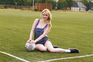 chica rubia con una pelota sentada en un campo de fútbol.