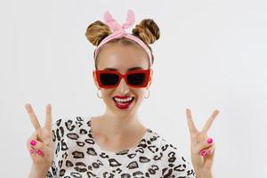 Close up fashion woman portrait isolated on white background. Happy girl face in sunglasses. Peace sign. Amazing and excited female with funny hairstyle. Selective focus photo