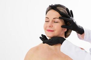 Hermosa mujer de mediana edad preparándose para el levantamiento de párpados, cirugía plástica, manos del médico con guantes negros señalan con el dedo a la cara en blanco. concepto de belleza, personas y salud. foto