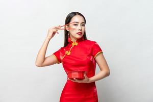 hermosa mujer asiática con vestido tradicional chino rojo qipao mirando una cámara sosteniendo una caja de comida y palillos en un estudio aislado de fondo gris foto