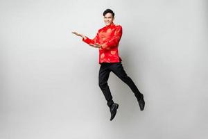 Jumping portrait of smiling handsome Asian man in oriental attire opening hands to empty space aside in light gray isolated studio background for chinese new year concept photo